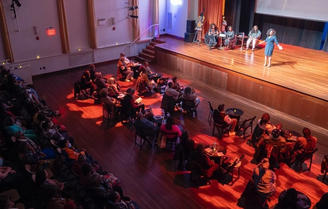 People attending a live performance on stage, with some seated and some standing, while an audience watches from seats and tables in a theater.