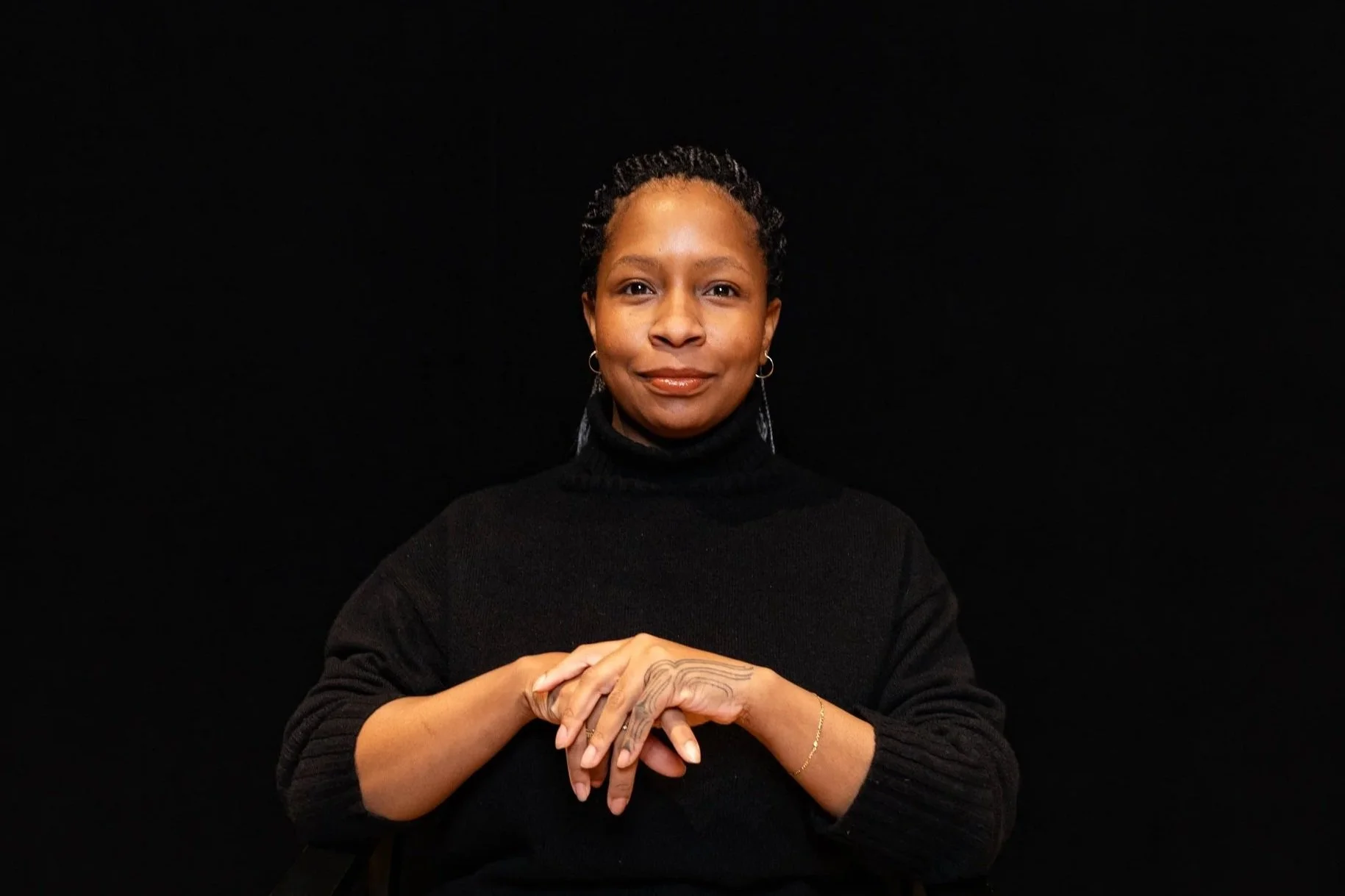 A woman with short, curly hair and dark skin, wearing a dark shirt, sitting in front of a large green leafy plant, with a blurred background of a window and outdoor scenery.