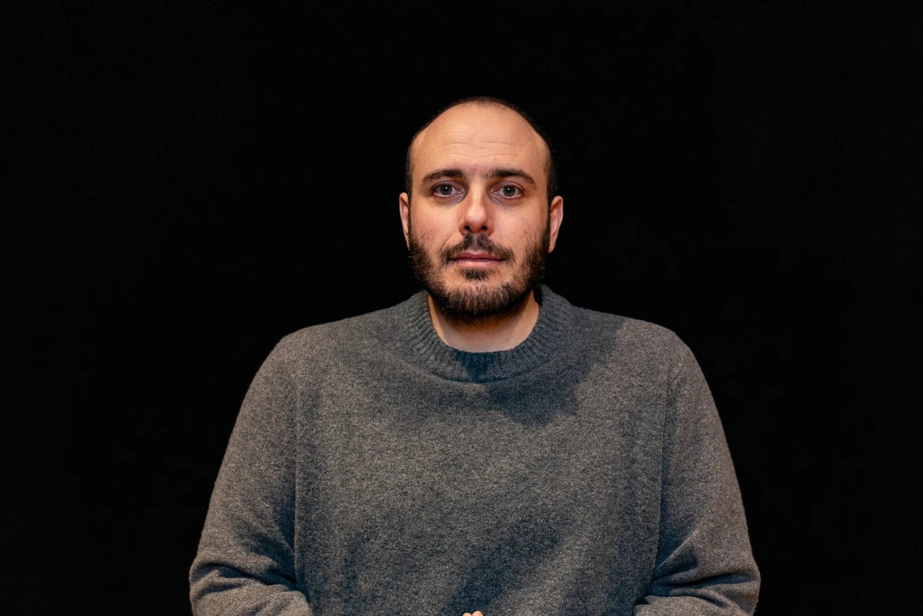 A man with a bald head and beard wearing a black suit, white shirt, and striped tie, standing against a neutral background.