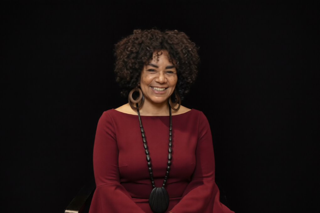 A smiling woman with curly black hair, wearing a black top, red and black beaded necklace, and hoop earrings, sitting indoors with wooden shelves in the background.