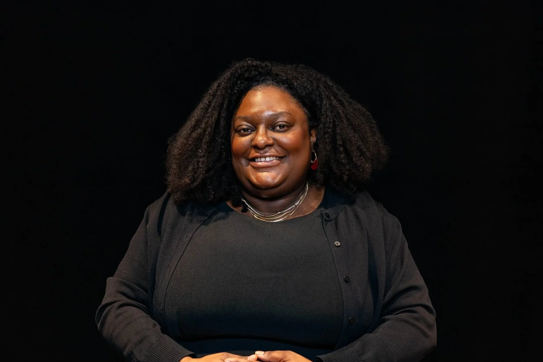 A woman with braided hair smiling wearing a black top and a gold chain necklace.