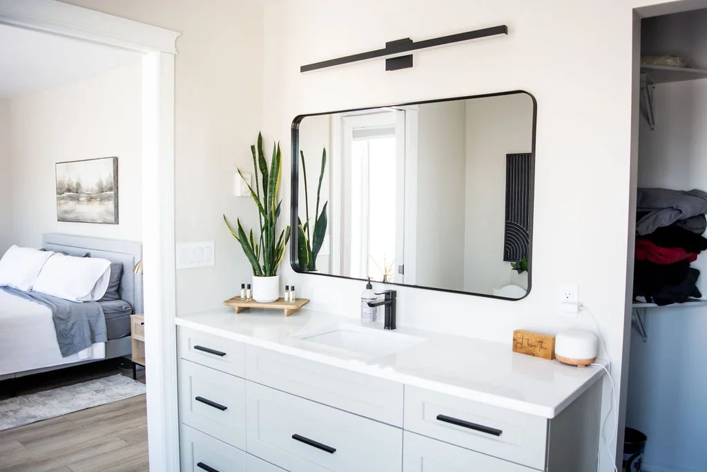 Modern white bathroom vanity with a large mirror, a tall green plant in a white pot, and a black faucet. Next to it, there is a closet with folded clothes. The reflection in the mirror shows a window and part of a bedroom with a bed and a landscape p