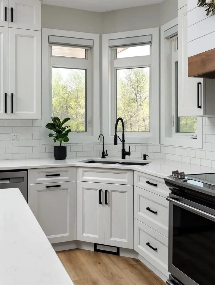 A modern kitchen with white cabinets, a black sink with a black faucet, three large windows with views of trees, a countertop plant, and black hardware on the cabinets. There is also a black oven visible.