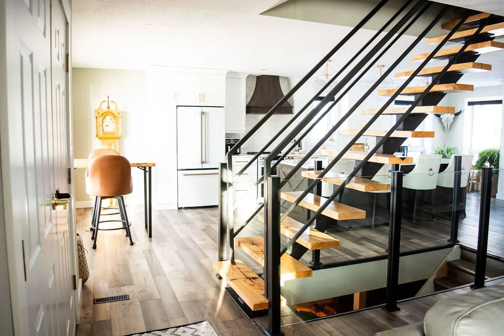 Interior view of a modern home with a staircase, kitchen, and sitting area, featuring light-colored walls, hardwood flooring, and contemporary furniture.