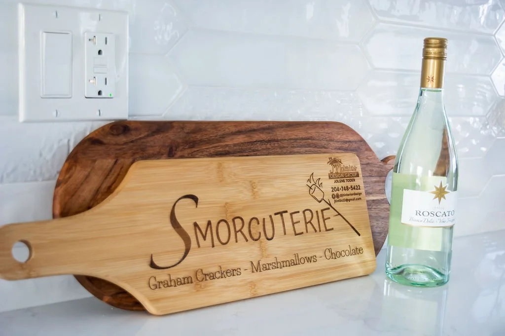A wooden serving board with engraved text for a store called Smorcuterie, advertising graham crackers, marshmallows, and chocolate, and a bottle of Starrosato sparkling wine on a kitchen countertop with white backsplash tiles and an electrical outlet