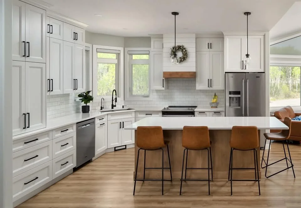 Modern kitchen with white cabinets, black handles, a large island with four brown chairs, a stainless steel refrigerator, and large windows letting in natural light.