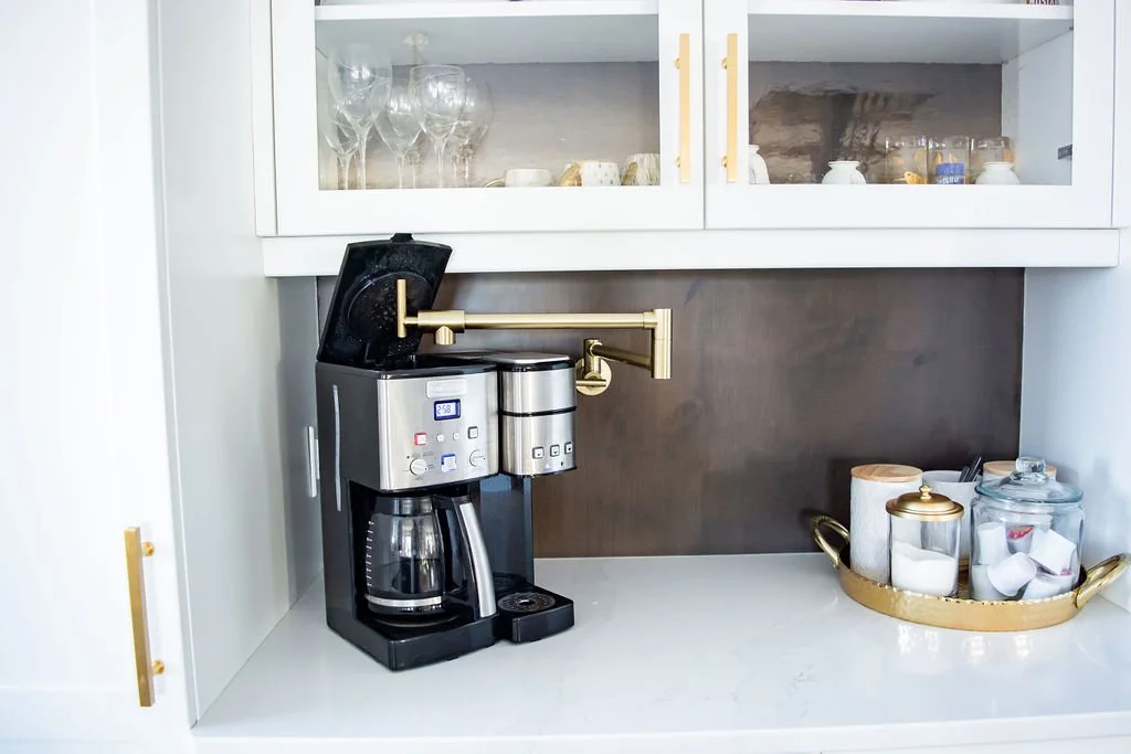 Coffee station with a drip coffee maker, glass jars, and cups in a white kitchen cabinet.