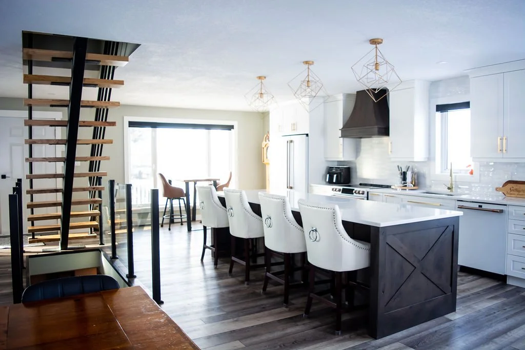 Modern kitchen with white cabinets, a large island with white countertop, gold hardware, and white cushioned barstools. Open dining area with a round table and chairs, large windows, and hanging pendant lights. Wooden staircase with open risers.