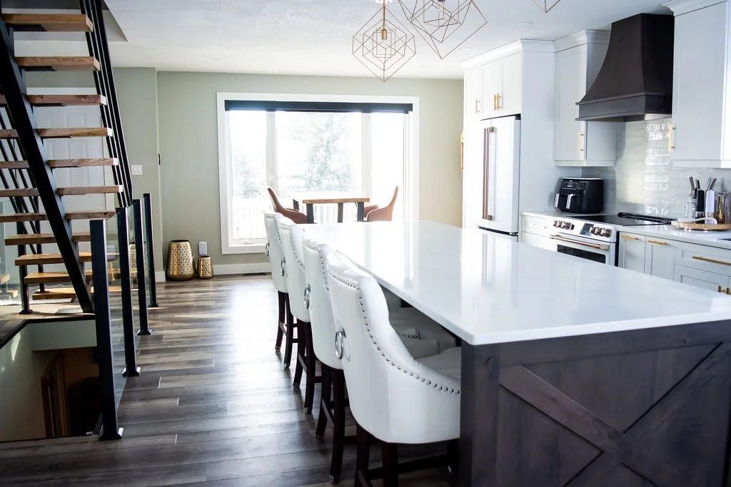 Modern kitchen with white cabinets, a large white island with white upholstered chairs, stainless steel appliances, and a dining area with a round table and four chairs near a window.