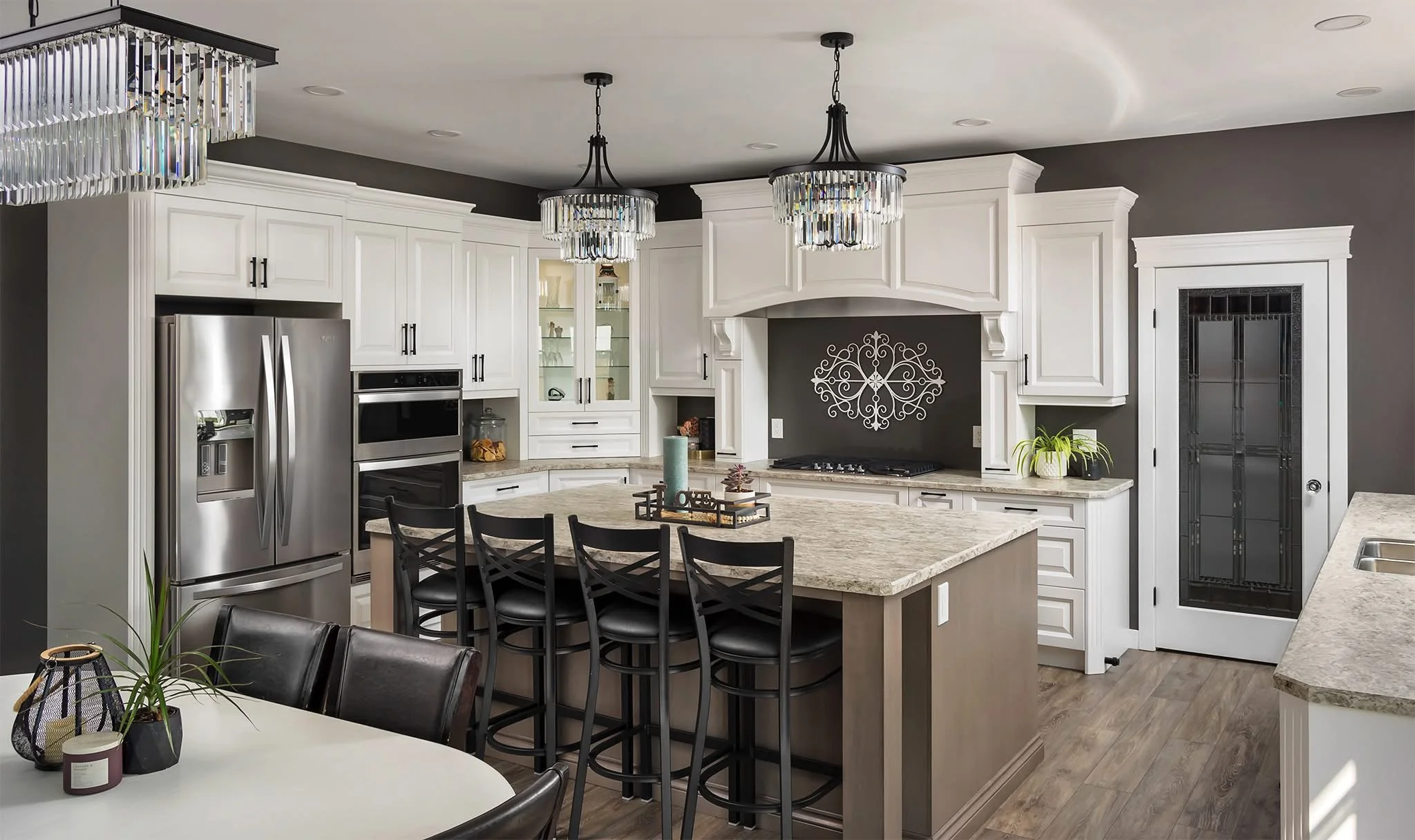 Modern kitchen with white cabinets, stainless steel refrigerator, black chairs at island, and decorative overhead lighting fixtures.