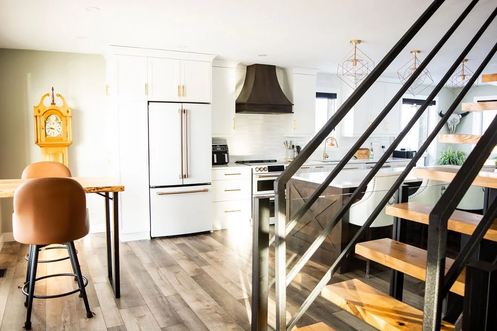 Modern kitchen with white cabinets, a black range hood, and a kitchen island with chairs under hanging geometric pendant lights. There is a wooden staircase with black metal railing and a wooden floor.