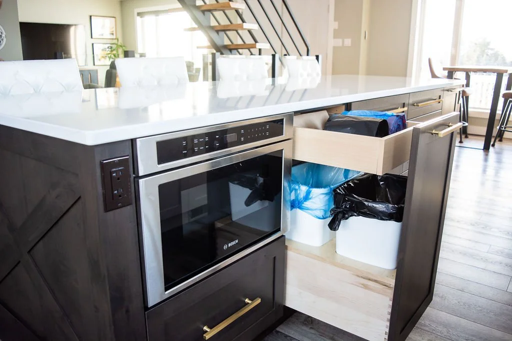 Open kitchen drawer filled with trash bags, trash cans, and cleaning supplies under white kitchen countertop with modern appliances and chairs in background.