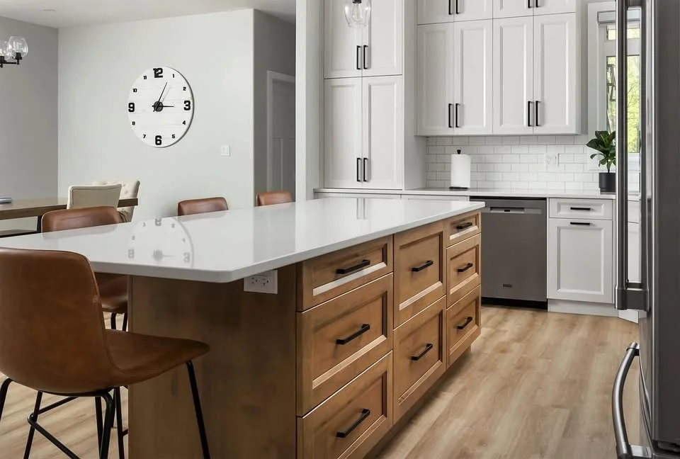 Modern kitchen with white cabinets, a large white island, and a wooden floor. There are leather chairs around the island and a clock on the wall.