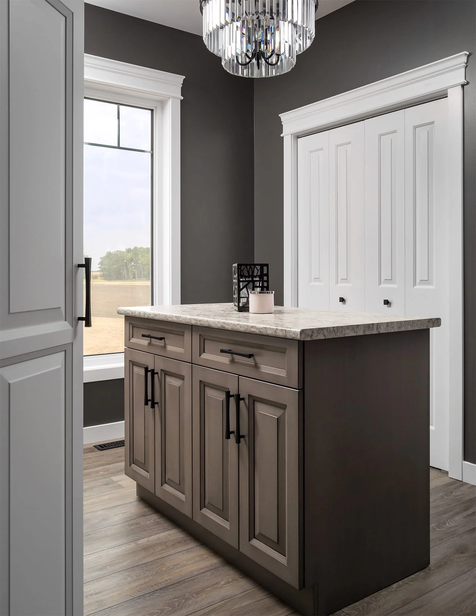 Modern laundry room with dark gray walls, white trim, a window, a light-colored wood floor, a gray cabinet with black handles, a marble countertop, a black decorative item, and a white closet with sliding doors.