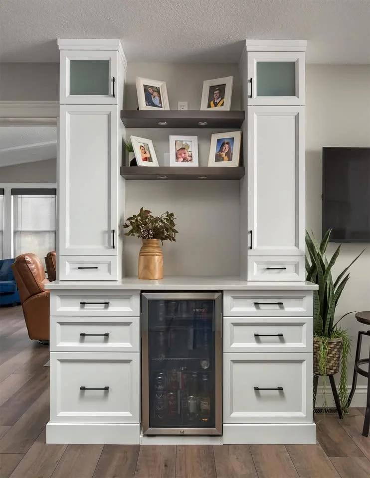 White kitchen cabinet with drawers, a small wine fridge, and two open shelves with framed photographs, a potted plant, and living room furniture visible in the background