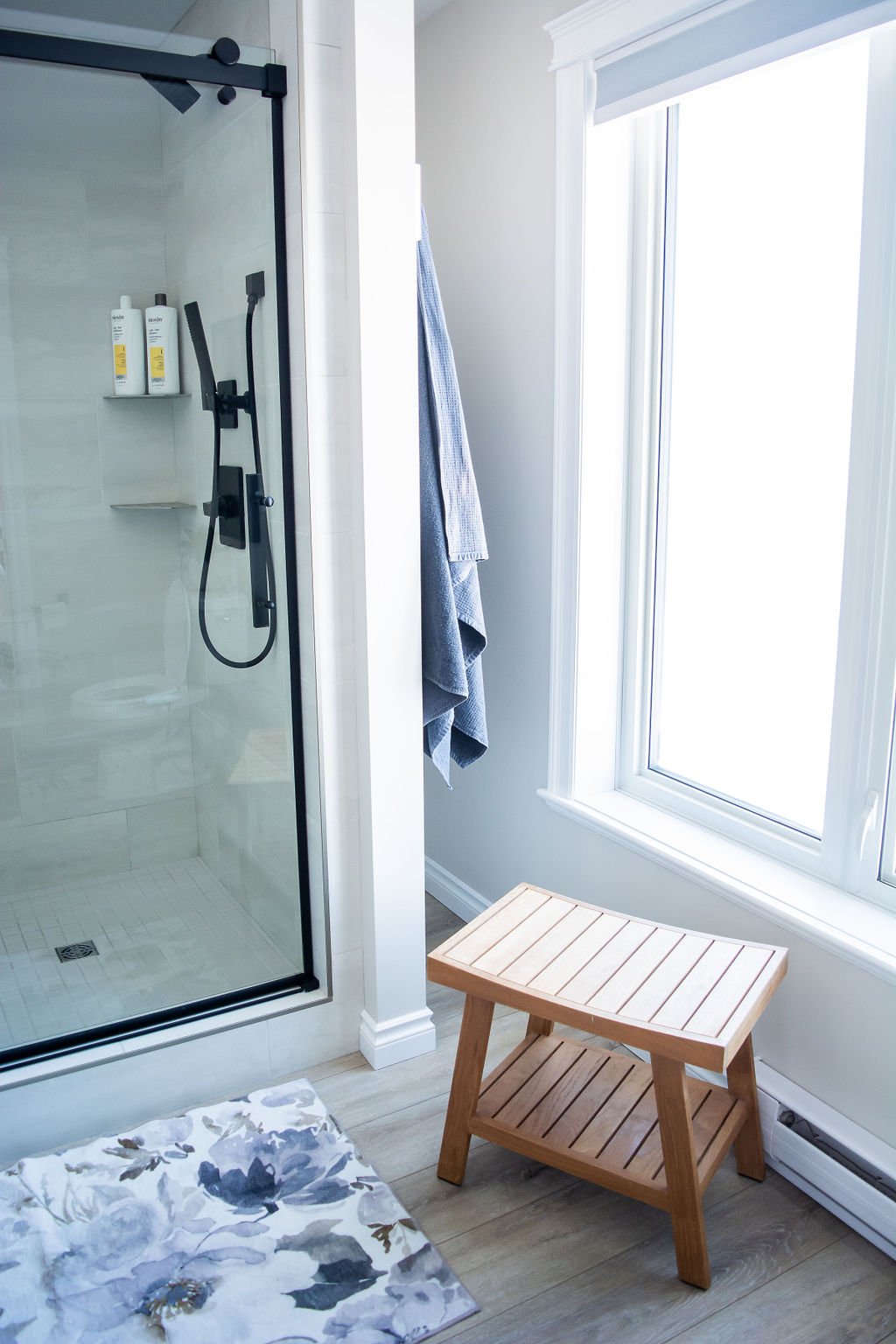 Shower with black frame and white tiles, two gray towels hanging on a hook, a wooden stool near a large window, and a floral rug on a light wood floor in a bathroom.