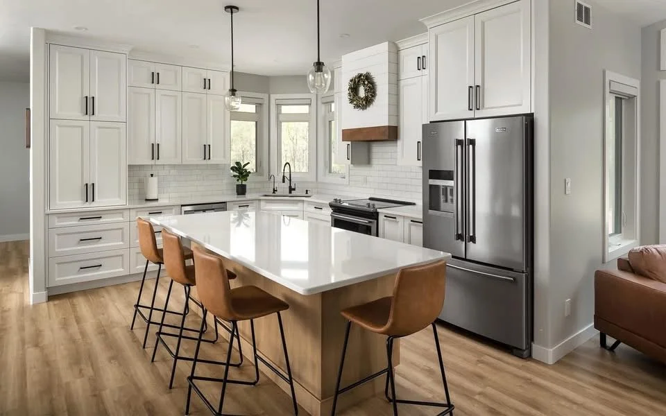 Modern kitchen with white cabinets, a large white island with brown bar stools, stainless steel refrigerator, black oven, and a window above the sink. Wooden flooring and a porch wreath above the stove.