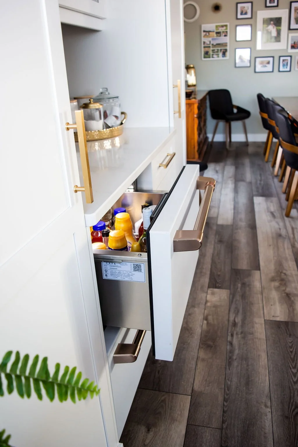 Open kitchen drawer filled with various snack bottles and condiments, part of a modern kitchen with wooden flooring and a dining area in the background.