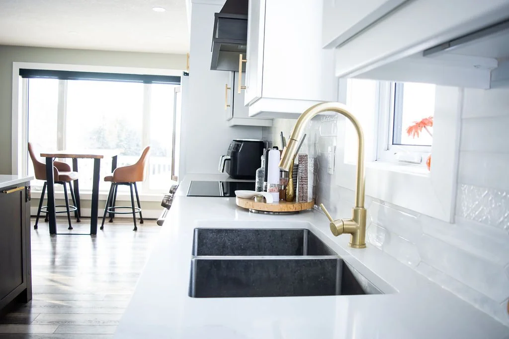 Modern kitchen with white countertops, black sink, gold faucet, and window overlooking a plant.