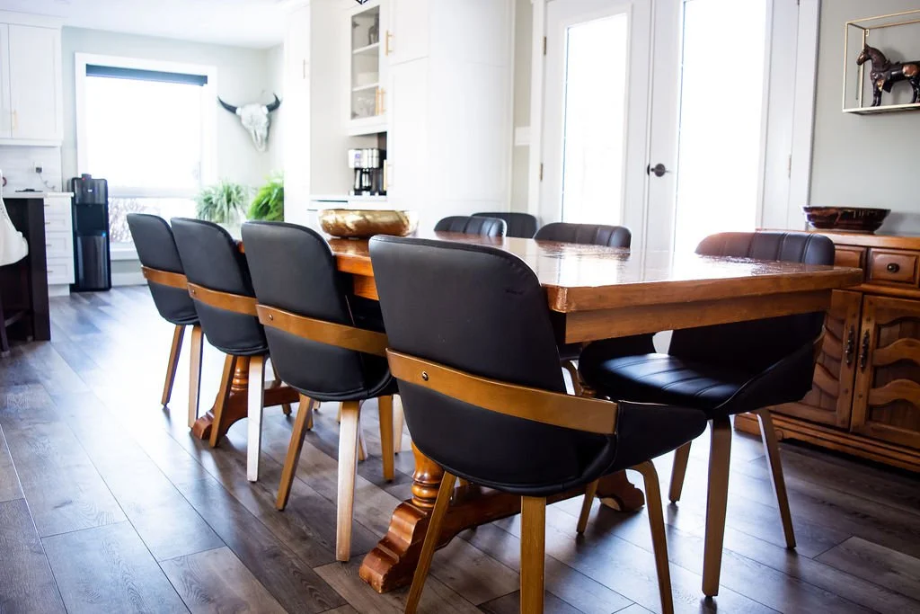 Dining room with a wooden table, six black and tan chairs, white walls, a window with a roller shade, and decorative items including a bull skull and a horse sculpture.
