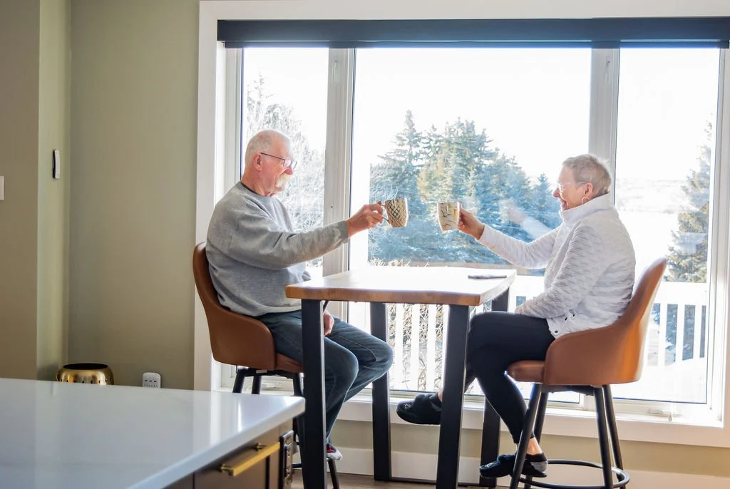 An elderly man and woman sitting at a small table by a large window, enjoying hot beverages and smiling at each other.