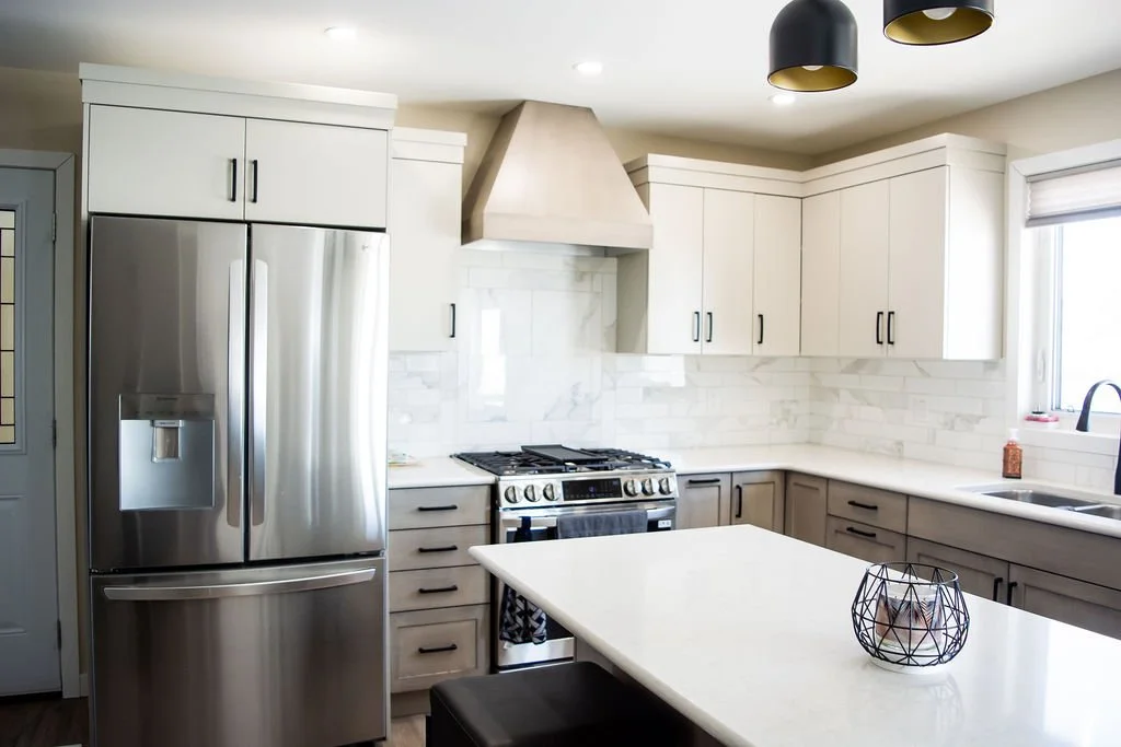 Modern kitchen with white and gray cabinetry, stainless steel refrigerator, built-in oven, white marble backsplash, window over sink, black pendant lights, and island with wire fruit basket.