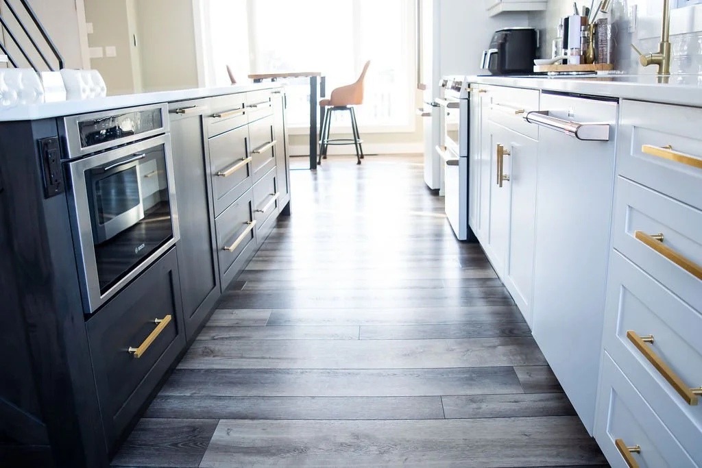 Modern kitchen with dark and light cabinets, gold hardware, stainless steel appliances, and wooden flooring, leading to a bright dining area with chairs and a table.