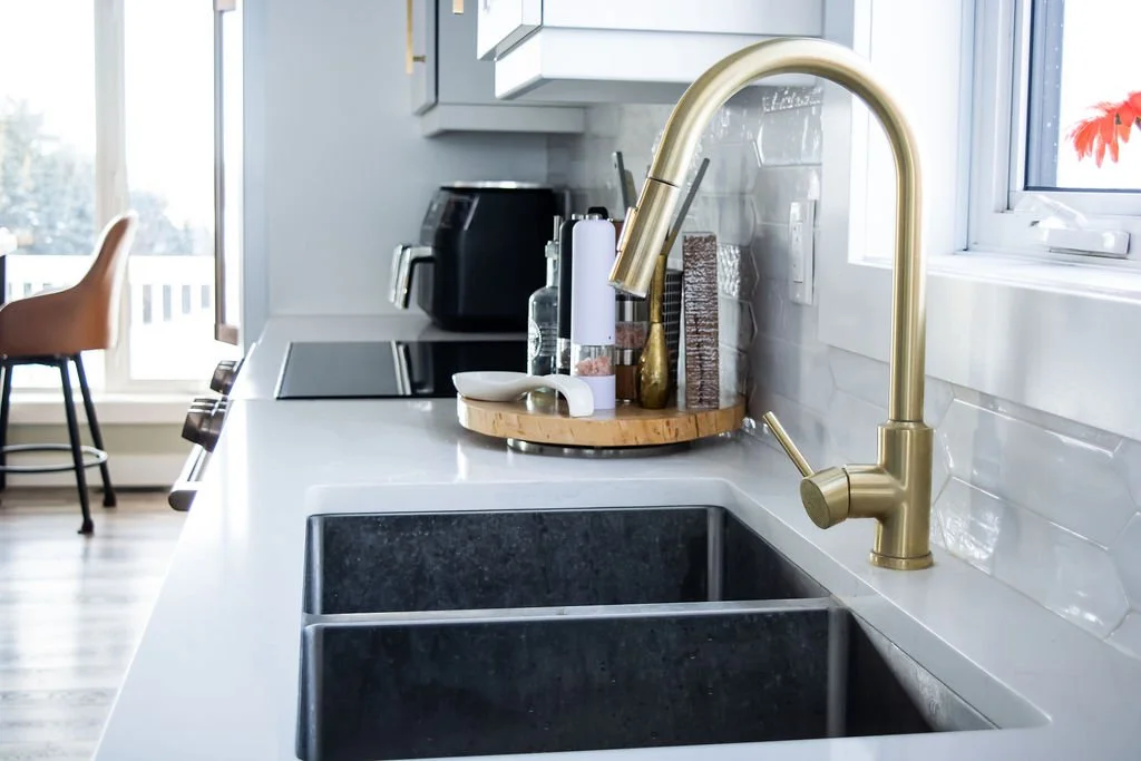 Modern kitchen sink with gold faucet, on white countertop with kitchen appliances and decor, with window and outdoor view.