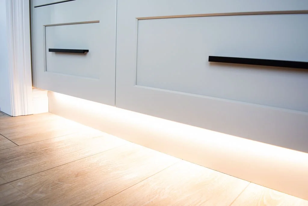 Close-up of a white cabinet with black handles and under-cabinet LED lighting on a wooden floor