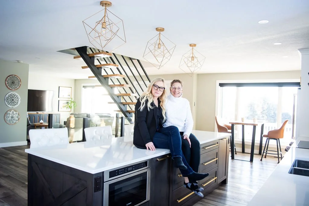 Two women sitting on a kitchen island in a modern, well-lit kitchen with a staircase, hanging geometric light fixtures, and large windows.