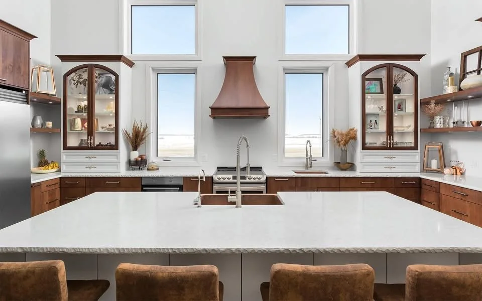 Modern kitchen with a large white island, brown cabinets, a copper range hood, stainless steel appliances, and open shelving with dishes and decor, natural light streaming through three large windows.