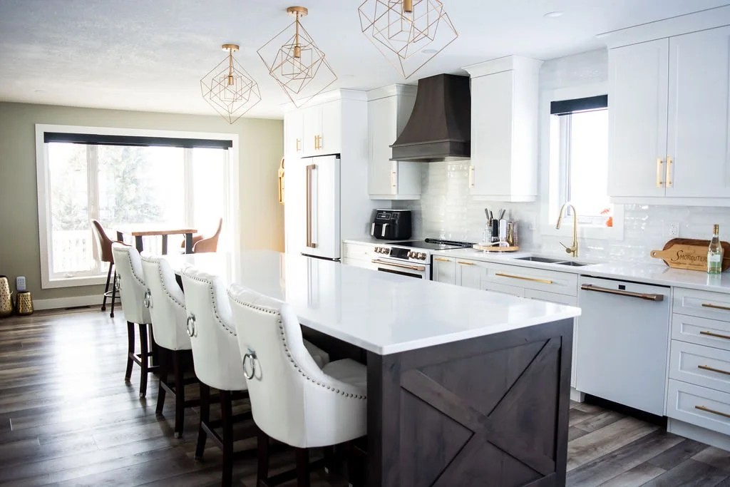 Modern kitchen with white cabinets, a large island with white countertop, black and white backsplash, and gold accents. There are hanging pendant lights above the island, a window over the sink, and a sliding glass door leading outside. Barstools lin