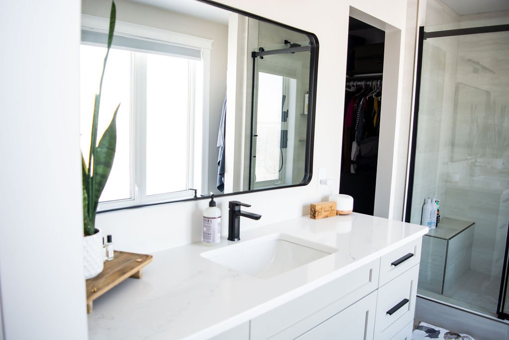 Modern bathroom with white vanity, black faucet, large mirror, potted plant, soap dispenser, and glass shower enclosure.
