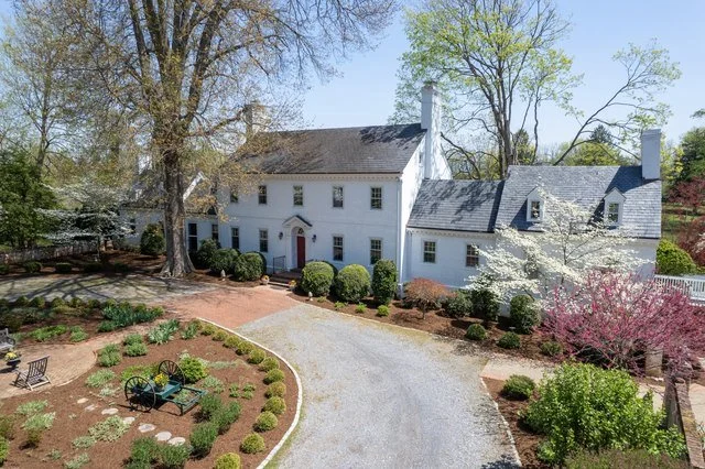 A large white historic house with a gray roof surrounded by a landscaped yard with a gravel driveway, shrubs, trees, and flowering plants.