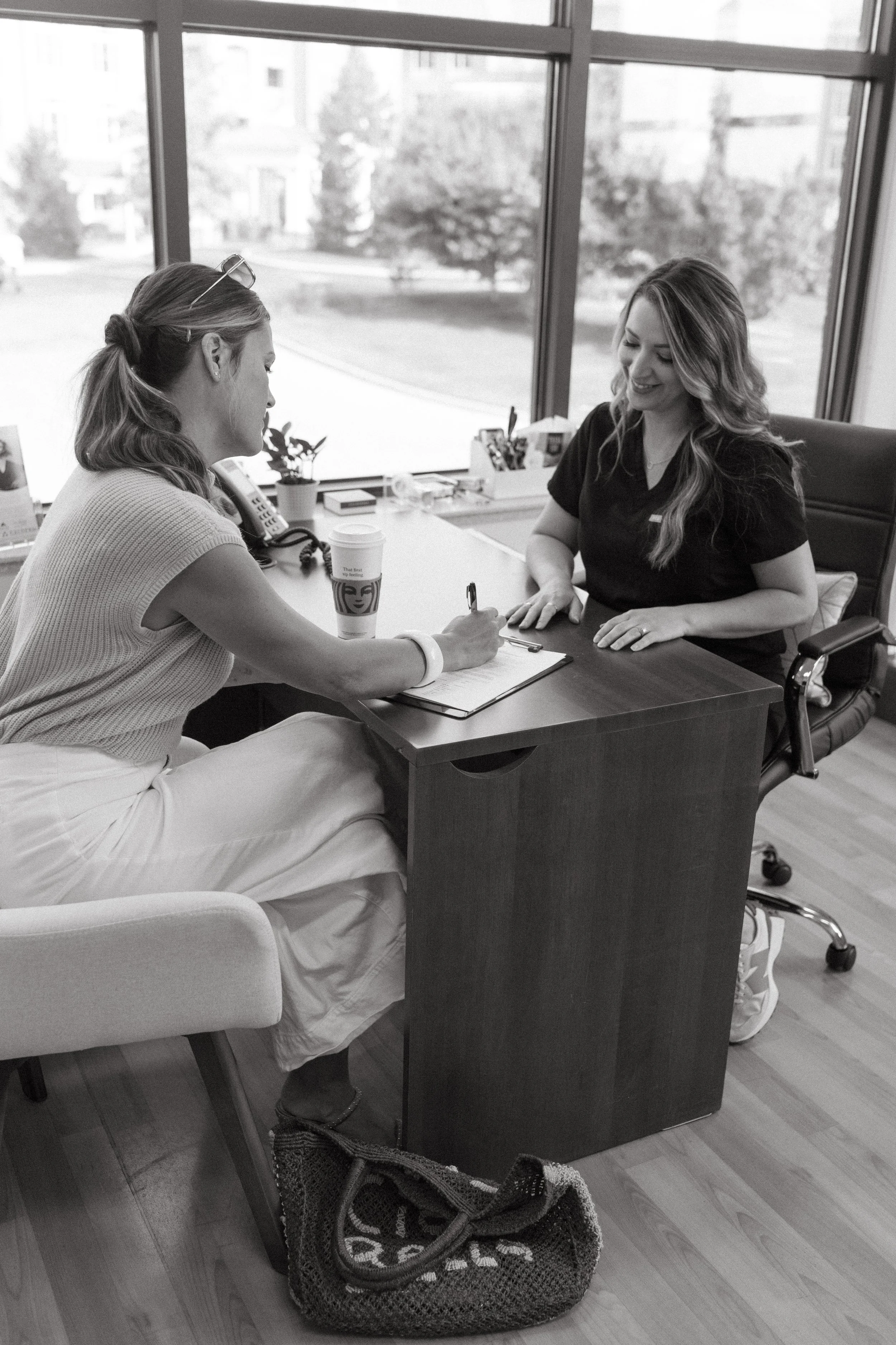 A woman in casual clothing sitting on a chair and signing a document for another woman in a black scrub top at an office desk. The woman on the chair has a coffee cup nearby, and there is a large window in the background showing trees and buildings.