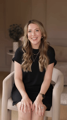 A woman with long wavy blonde hair, wearing a black dress, sitting in a beige armchair, smiling in a cozy room with neutral decor.