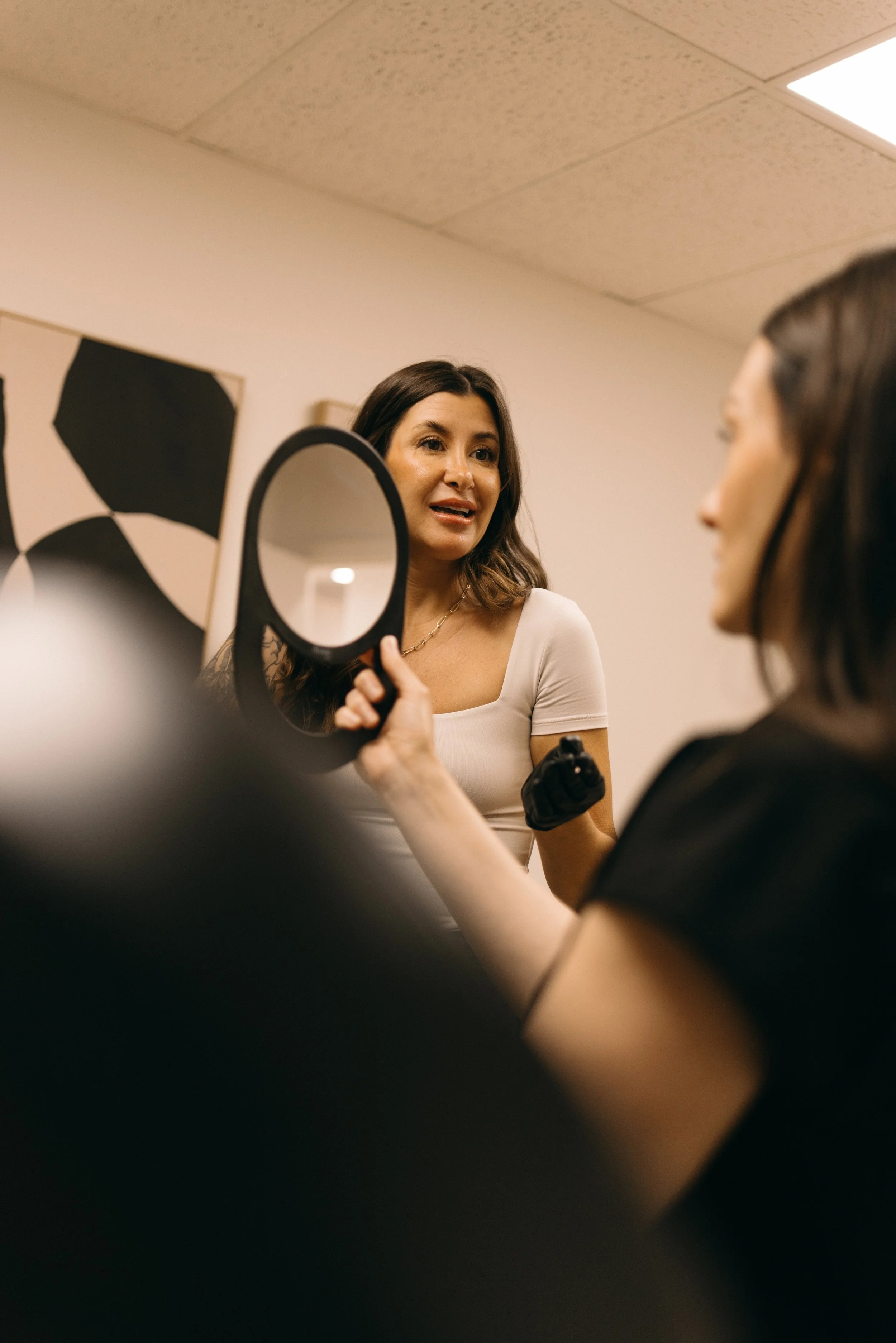 A woman with dark wavy hair is looking into a mirror held by another woman with black hair. The woman holding the mirror is wearing a black shirt, and the woman in front of her is wearing a light-colored top. The setting appears to be indoors, possibly a salon or dressing room.