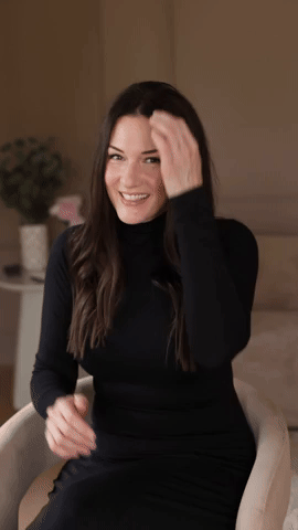 Smiling woman with long brown hair wearing a black turtleneck sitting in a room.