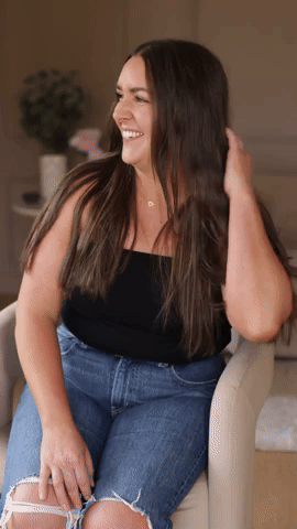Woman with long dark hair sitting on a chair, smiling, wearing a black top and ripped jeans, in a cozy indoor setting.