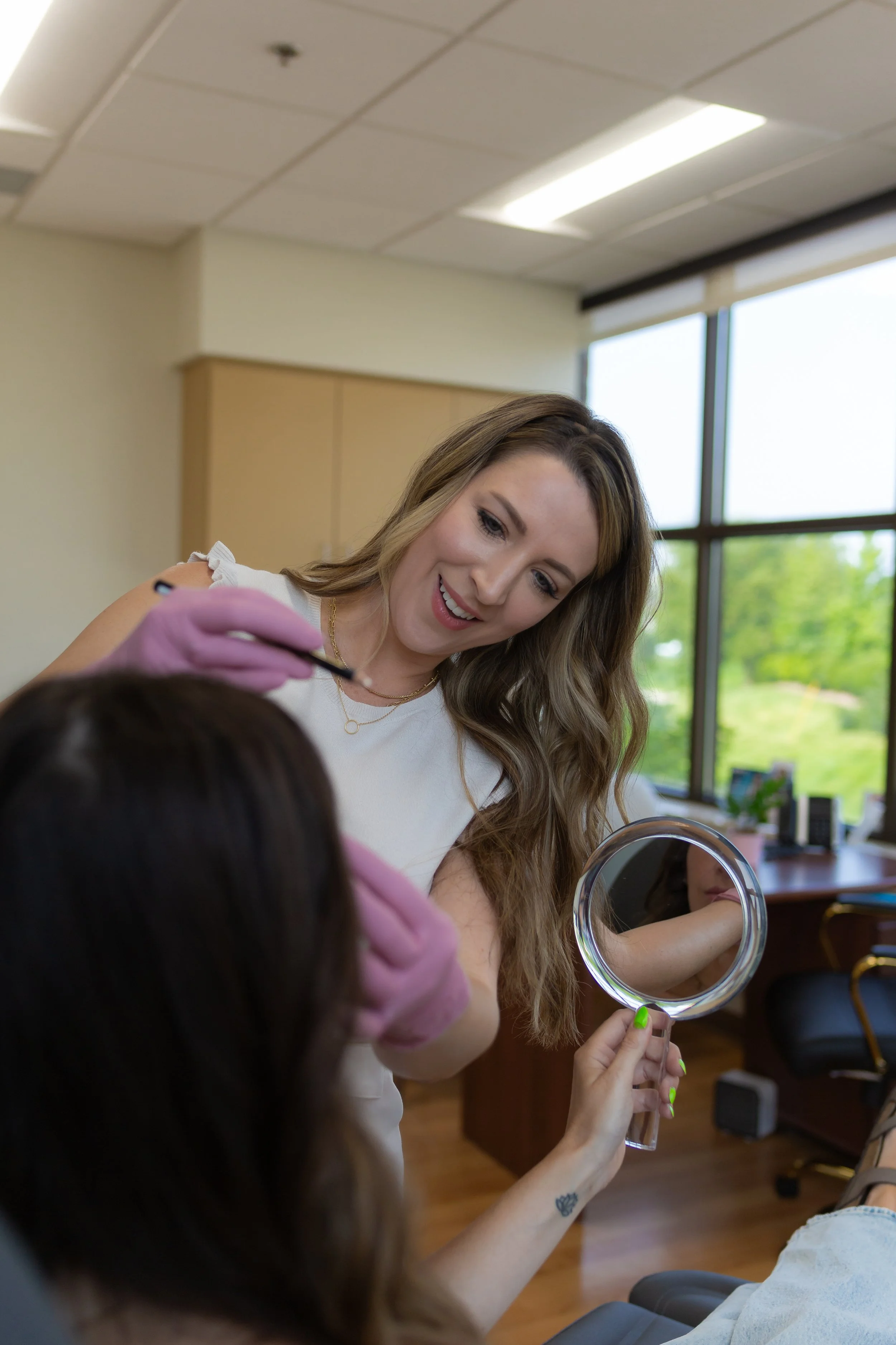 A woman with long, wavy hair and wearing pink gloves is applying makeup or performing beauty treatment on another woman who is holding a handheld mirror in an office setting with large windows showing green trees outside.