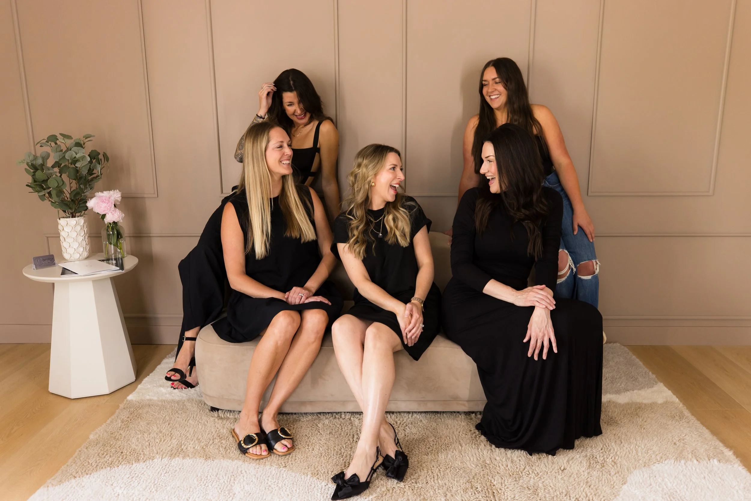 Six women sitting and standing together in a cozy room, smiling and laughing. One woman is sitting on a beige couch, three women are sitting on the floor or armrest, and two women are standing behind them. There is a small white side table with flowers and a notebook on it, next to the couch.