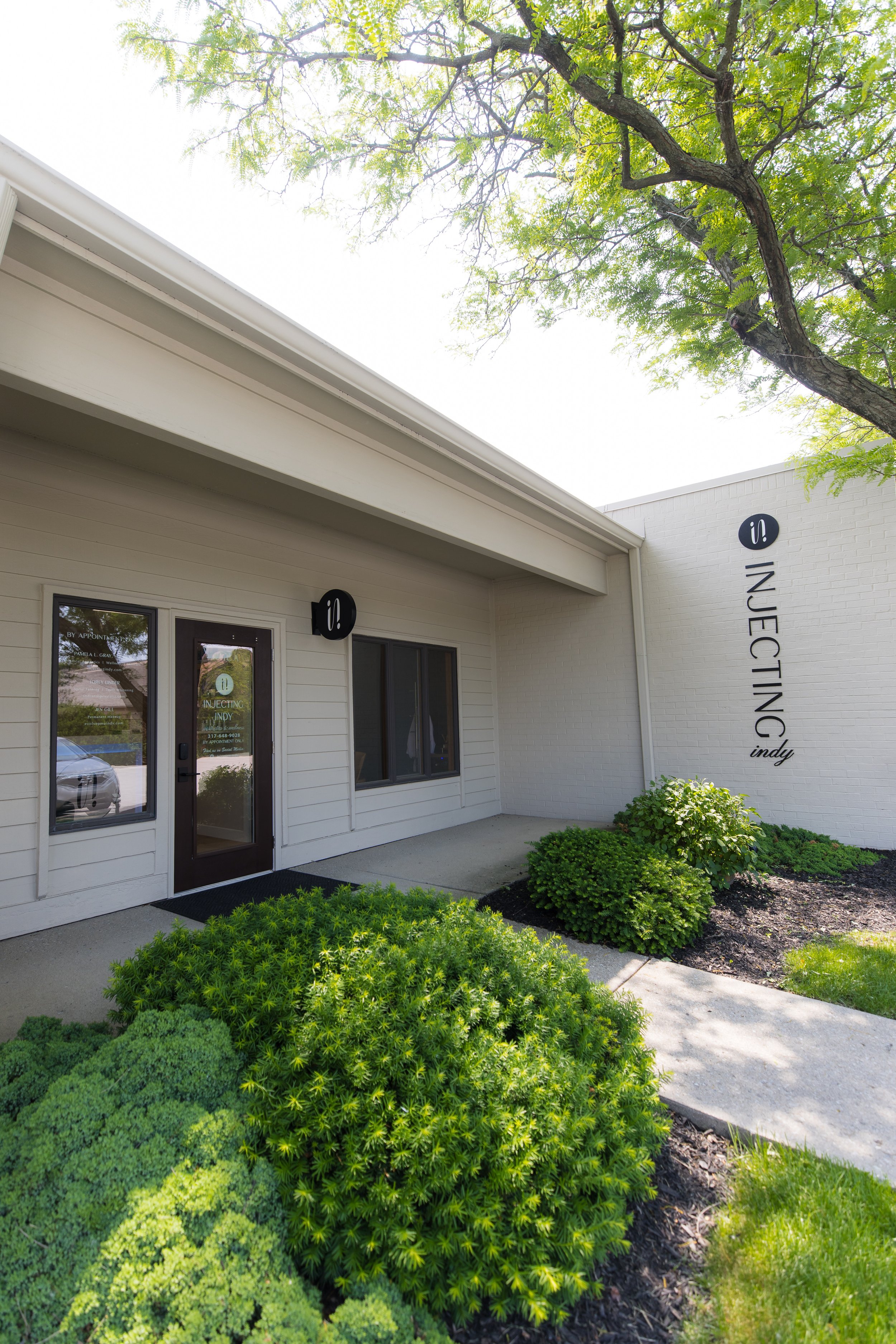 Exterior view of Injecting Indy clinic with white walls, black signage, front door, and landscaped bushes under a tree.