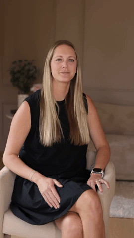 A woman with long blonde hair, wearing a black sleeveless dress, sitting on a beige armchair in a living room with beige walls and a plant in the background.