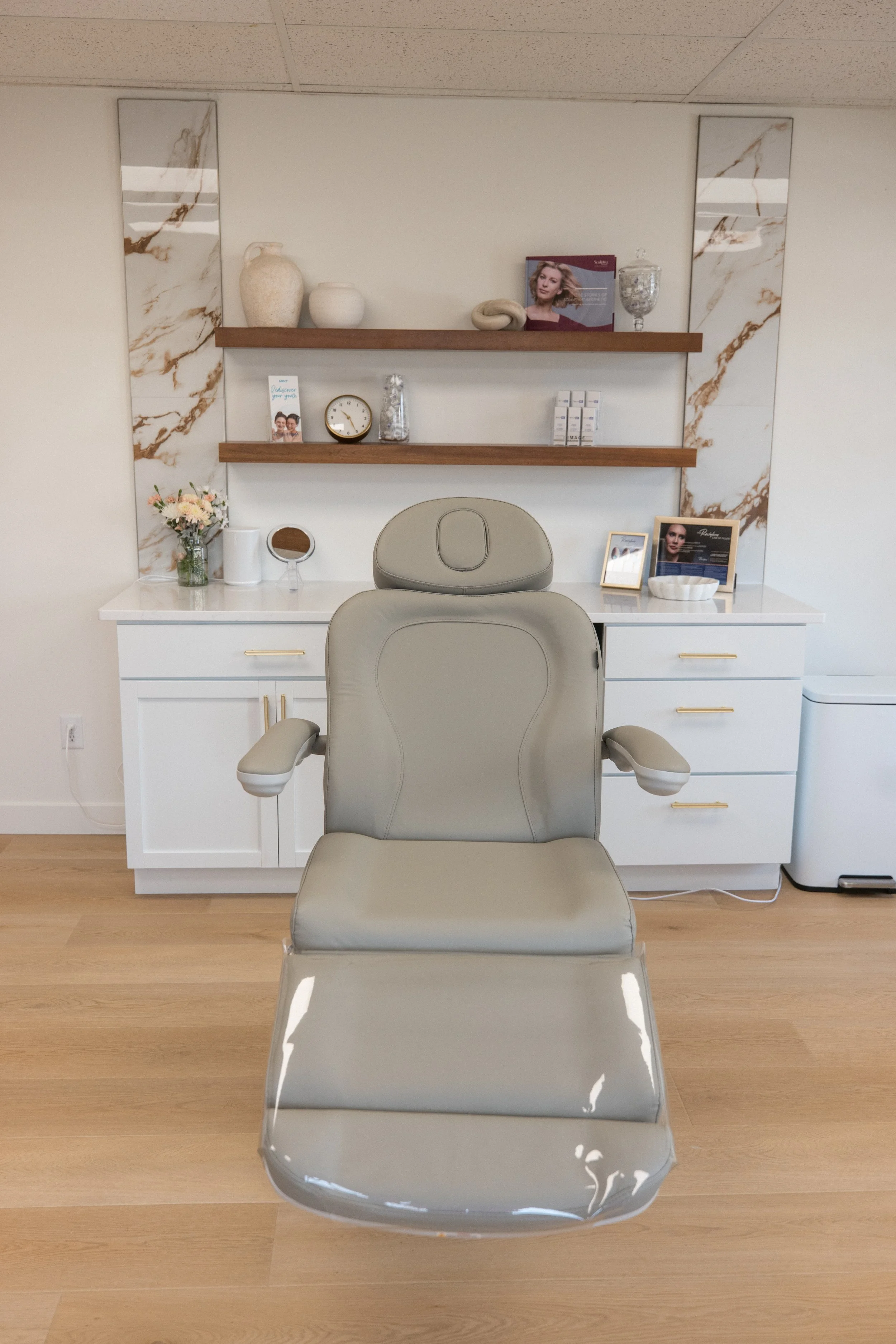 Medical or consultation room with a beige reclining chair centered in front of a white cabinet with gold handles, and two wooden shelves with decorative items above the cabinet.