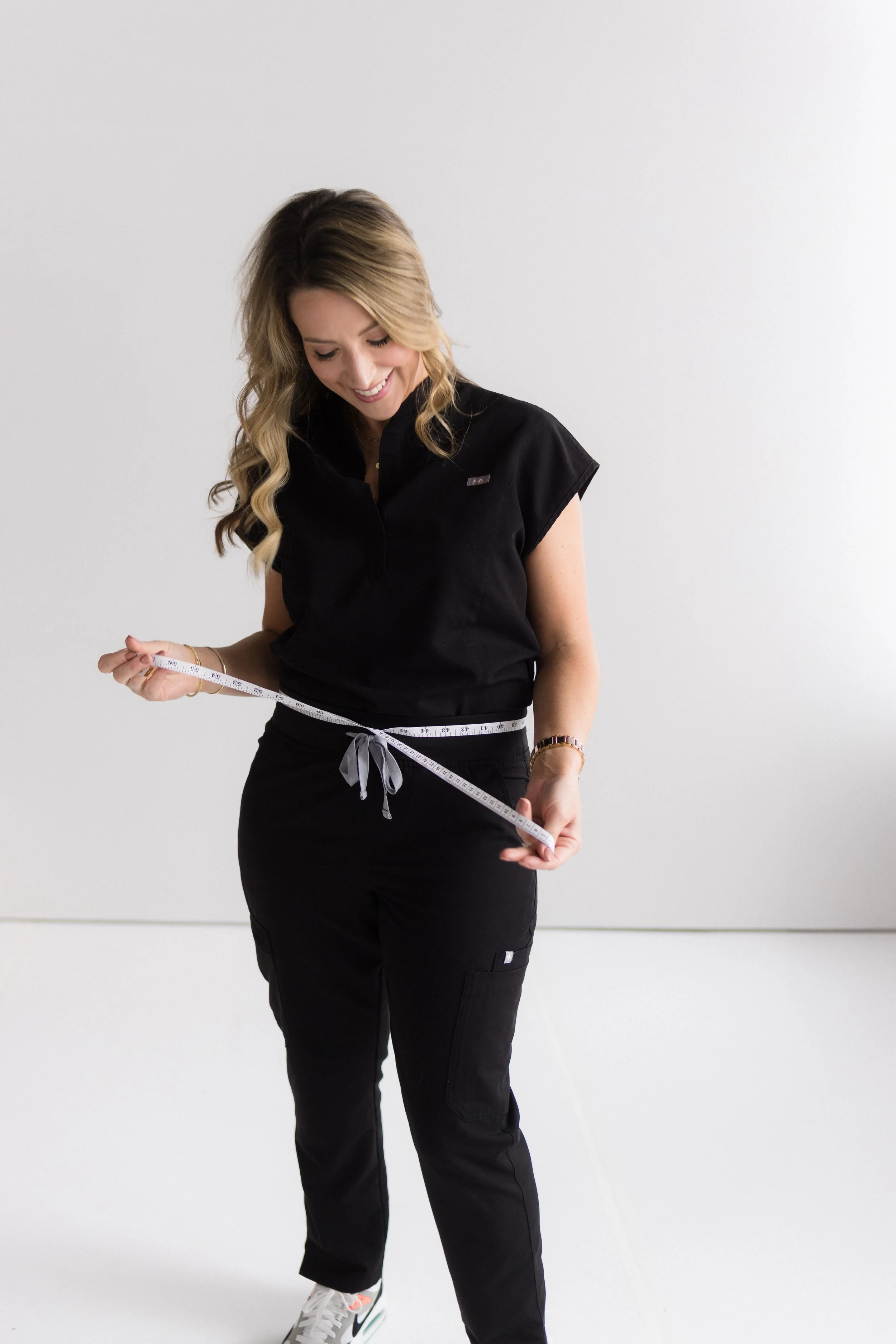 Woman smiling and holding a measuring tape around her waist, wearing black athletic clothing and sneakers.