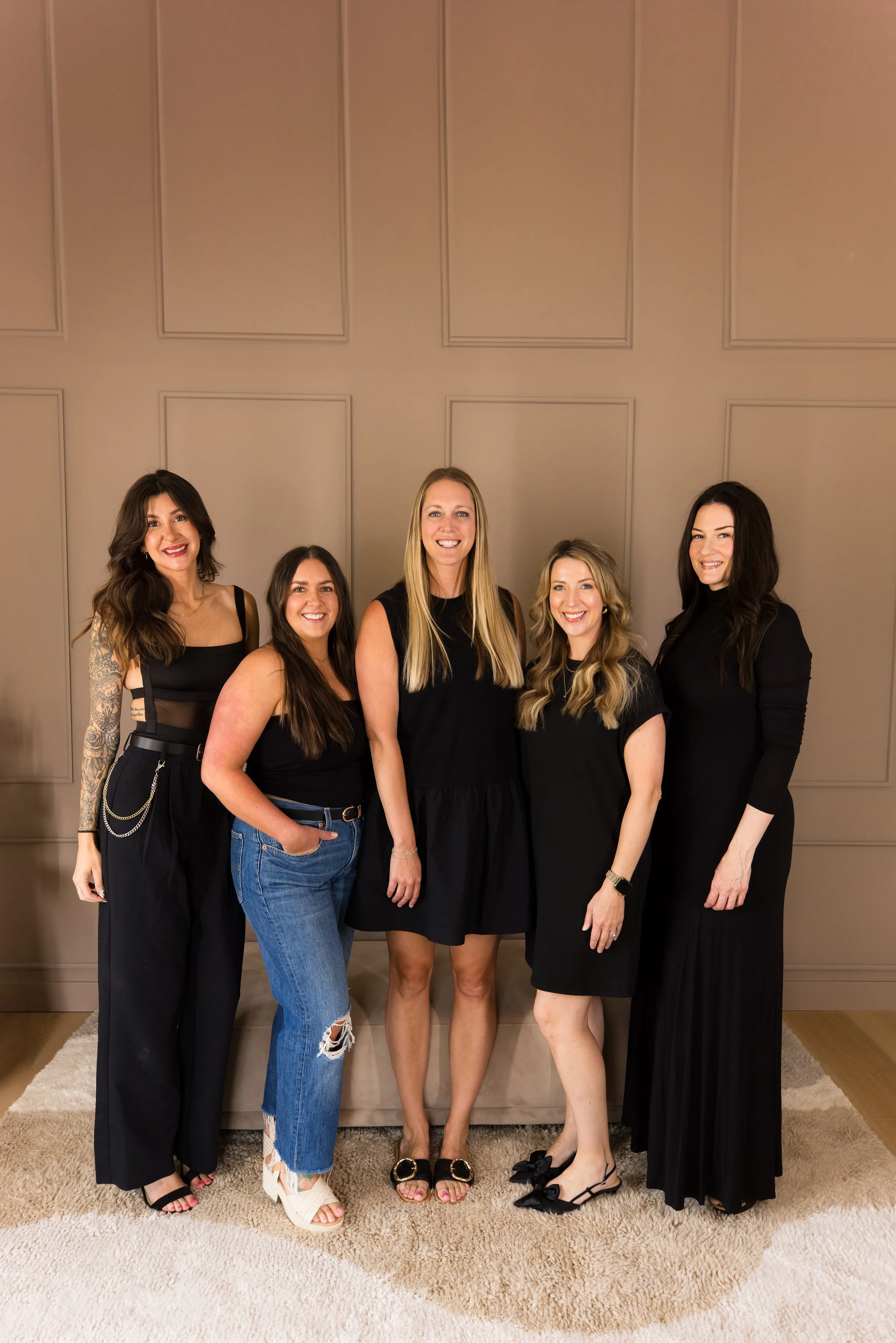 A group of five women standing together indoors in front of a beige wall with decorative paneling, smiling for the camera.