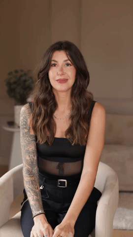 A woman with long, wavy dark hair, wearing a sleeveless black top with a sheer midsection, sitting on a beige armchair in a living room with a plant in the background.