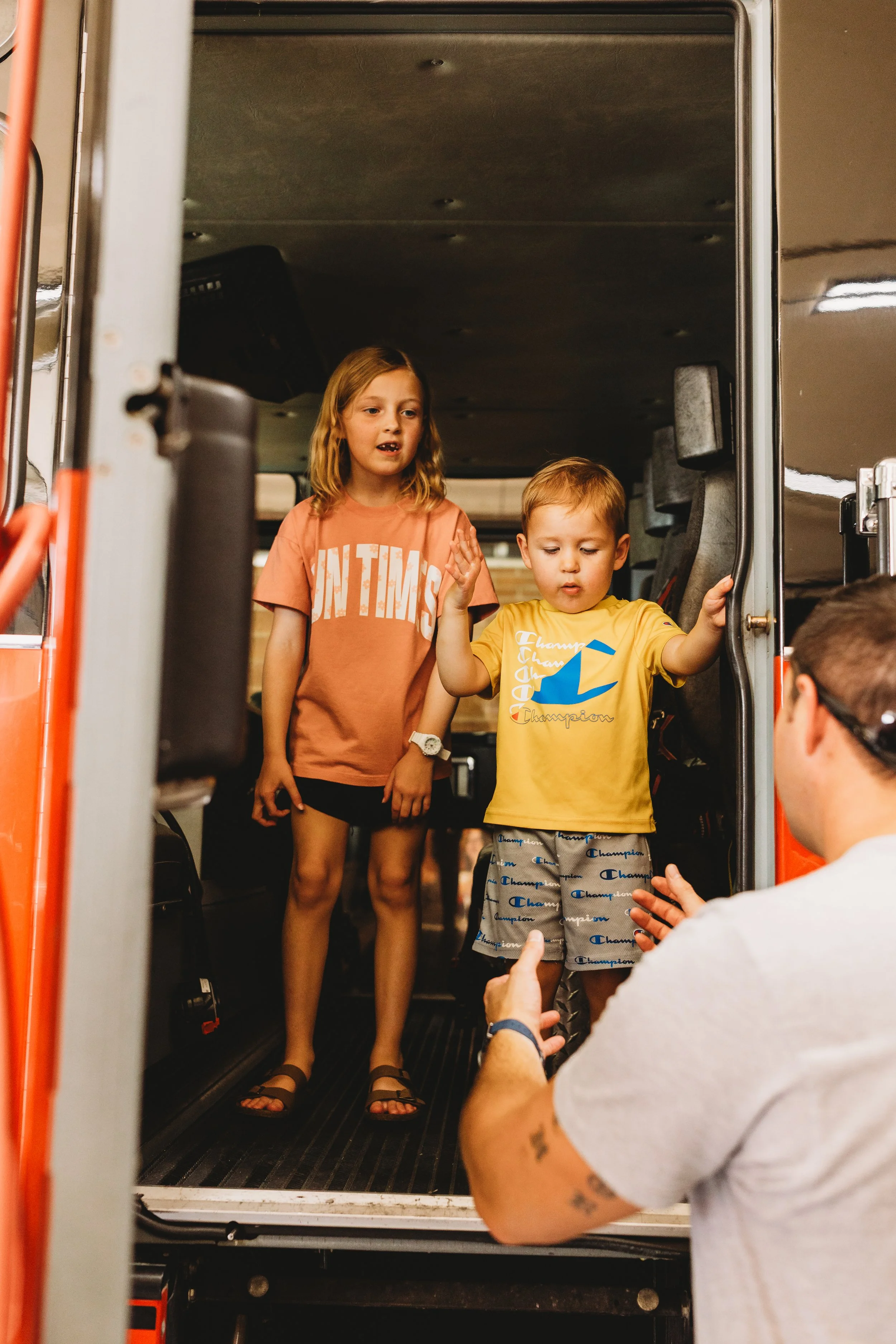 Two children, a girl and a boy, standing in the doorway of a fire truck, with a man talking to them.