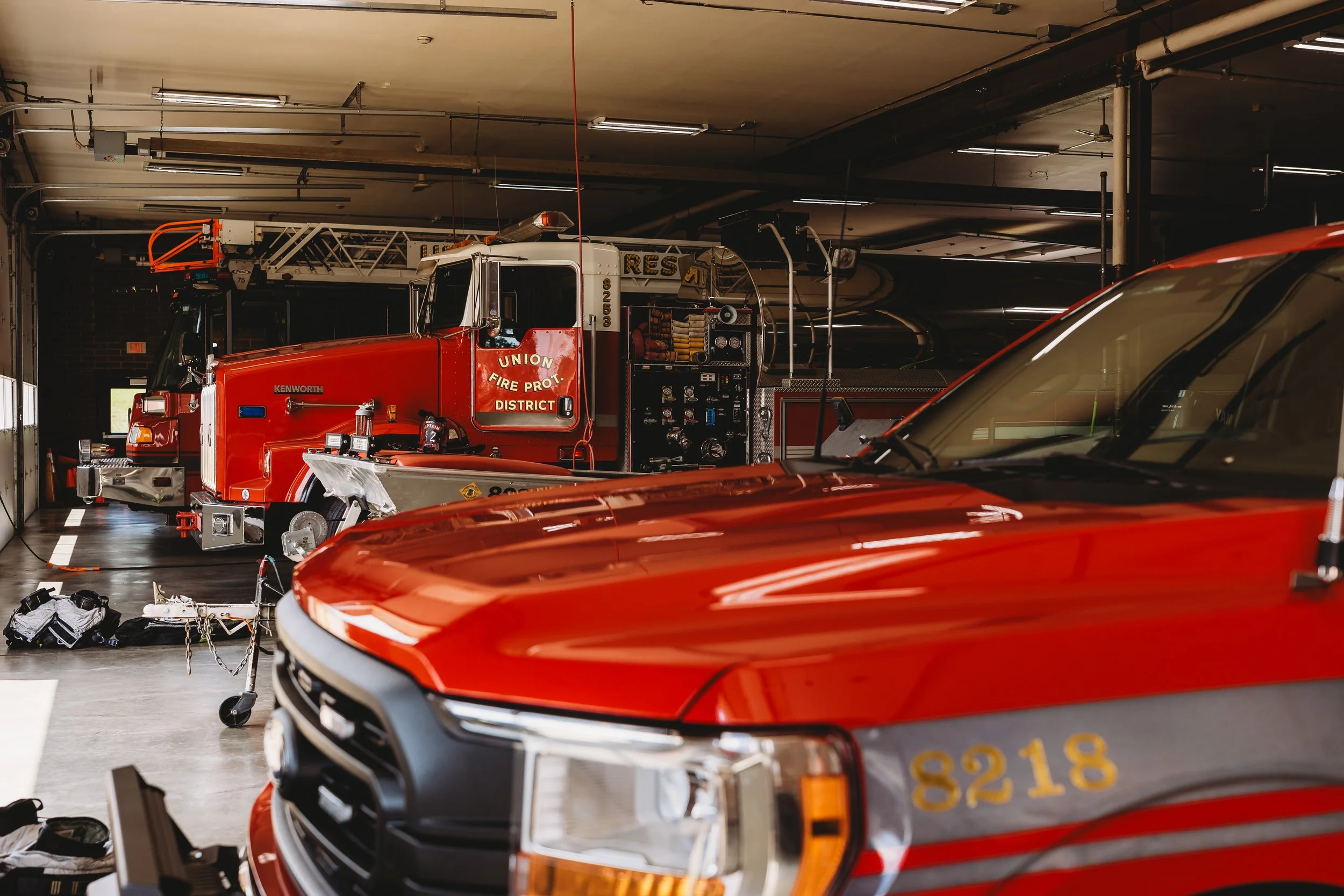 Inside a fire station garage with red fire trucks, including one labeled 'Union Fire Protection District.' Emergency equipment and tools are visible on the trucks and on the floor.