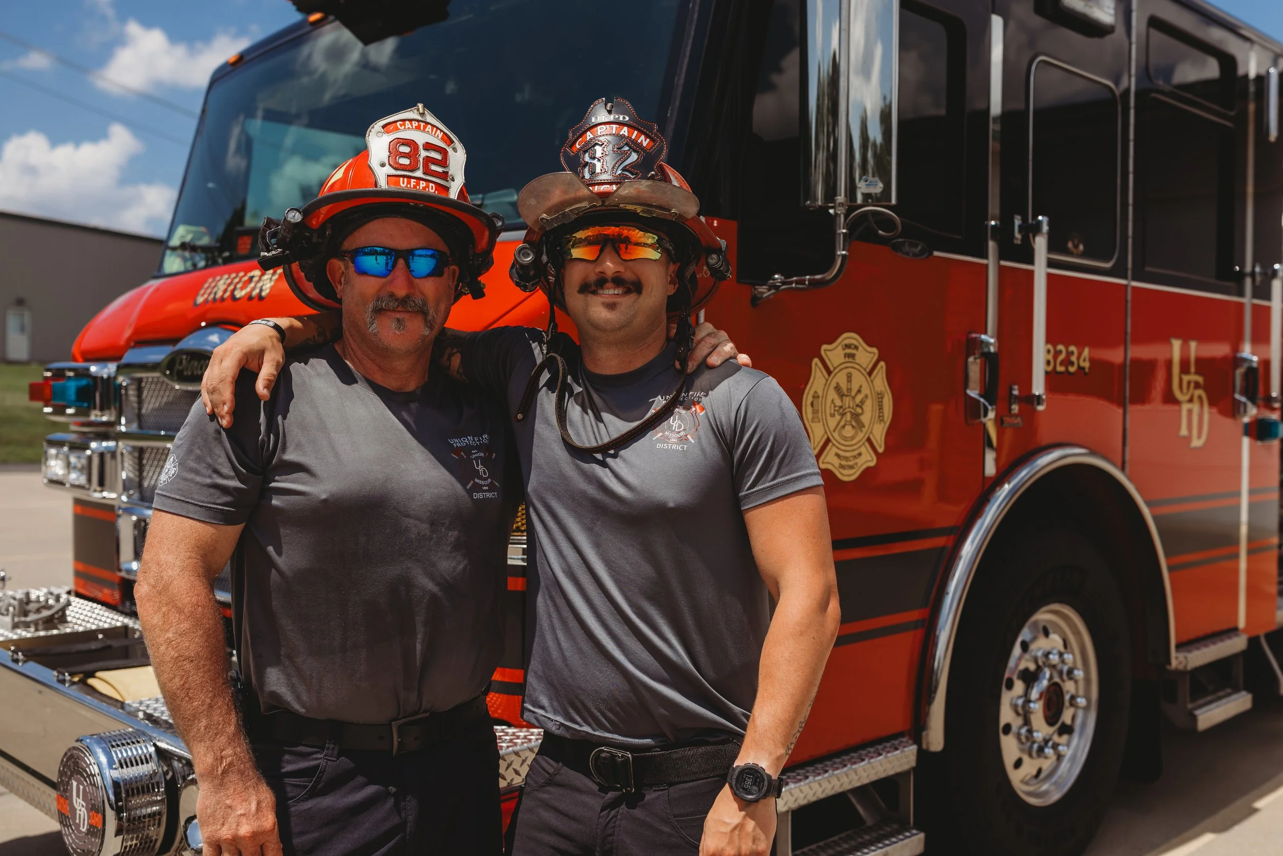 Two firefighters in uniform with helmets and sunglasses standing in front of a red fire truck, smiling and posing with their arms around each other.